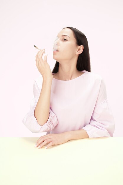スタジオのテーブルに座ってタバコを吸う若い女性。トレンディな色。コピースペースとミニマリズムスタイルの白人の女の子の肖像画