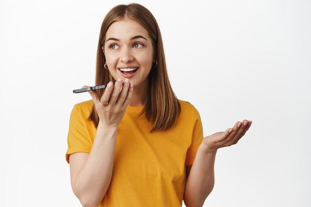 Free photo young woman on speakerphone, girl record message, voice call on smartphone, holding mobile near lips, translating words with application, standing against white background