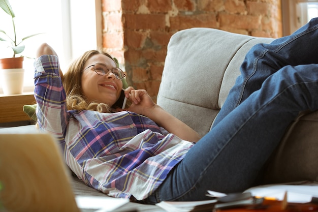 若い女性は、自宅のソファに横たわって電話で話します。