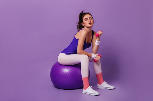 Молодая женщина в спортивном боди и розовых носках сидит на фитболе и пожимает руки гантелями на фиолетовой стене