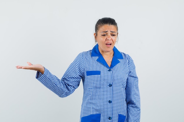 Young woman spreading palm as holding something in blue gingham pajama shirt and looking dismal , front view.