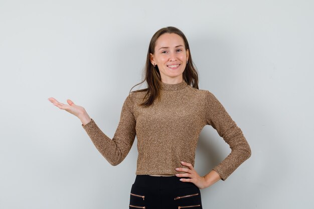 Young woman spreading palm aside in golden blouse and looking merry , front view.