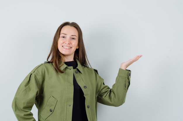 Young woman spreading palm aside in green jacket and looking pleased. front view.