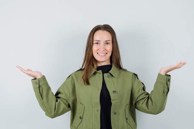 Young woman spreading palms aside in green jacket and looking cheery. front view.