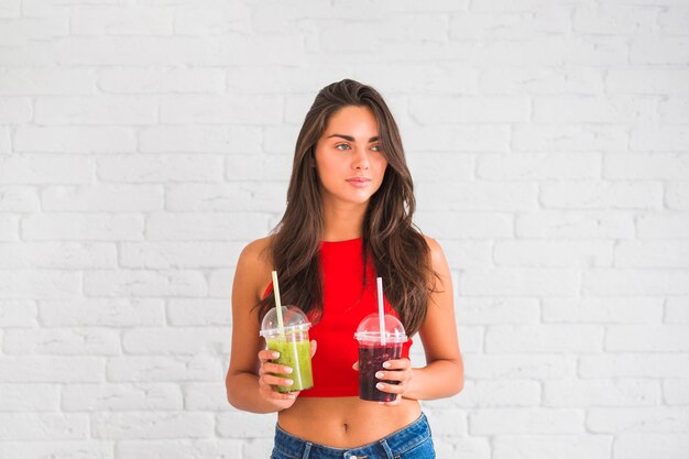 Young woman standing against wall holding smoothies in transparent plastic cup