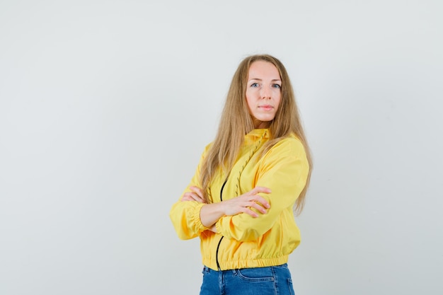 黄色のボンバージャケットとブルージーンズで腕を組んで立っている若い女性は、エレガントな正面図を探しています。
