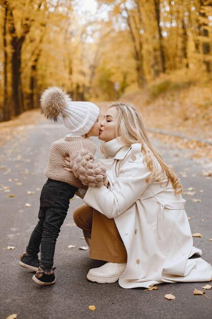 Young woman standing in autumn forest. Blonde woman kissing her daughter. Girl wearing beige sweater and white hat, mother wearing white coat.