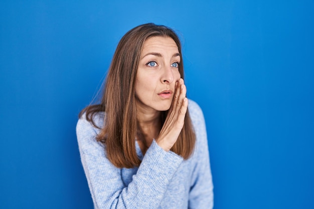 Foto gratuita giovane donna in piedi su sfondo blu mano sulla bocca che racconta voci segrete, sussurrando conversazioni maliziose