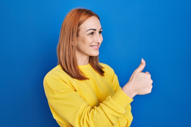 Young woman standing over blue background looking proud, smiling doing thumbs up gesture to the side