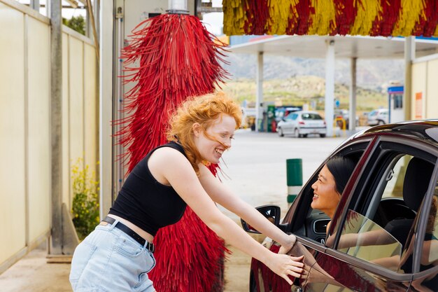 Young woman standing at car wash and smiling to Asian female looking out of car window 