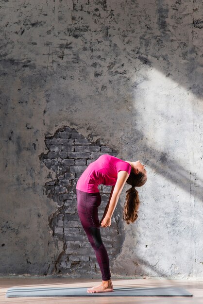 Young woman standing on exercise mat bending backward against an old wall