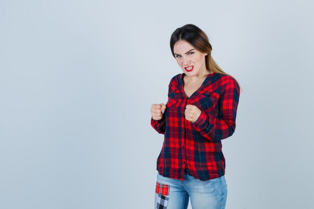 Young woman standing in fight pose in checked shirt, jeans and looking angry. front view.