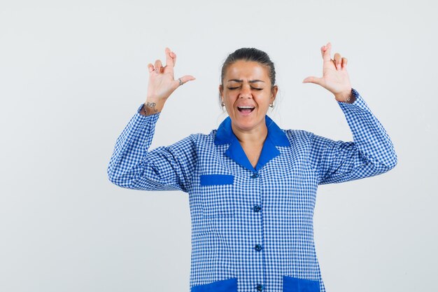 Young woman standing fingers crossed while closing eyes in blue gingham pajama shirt and looking pretty. front view.