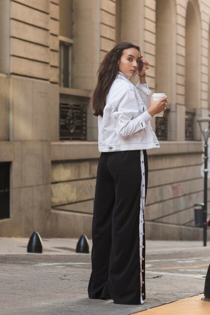 Young woman standing in front of building holding takeaway coffee cup looking over shoulder