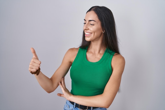 Young woman standing over isolated background looking proud, smiling doing thumbs up gesture to the side