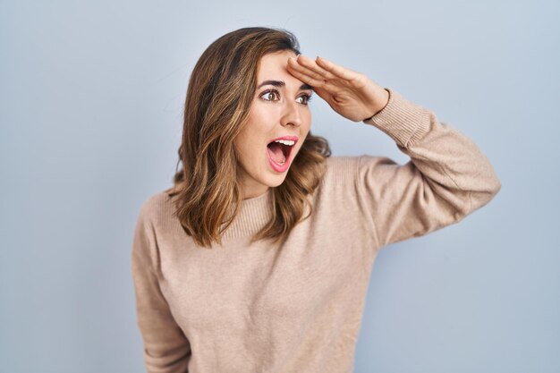 Young woman standing over isolated background very happy and smiling looking far away with hand over head searching concept