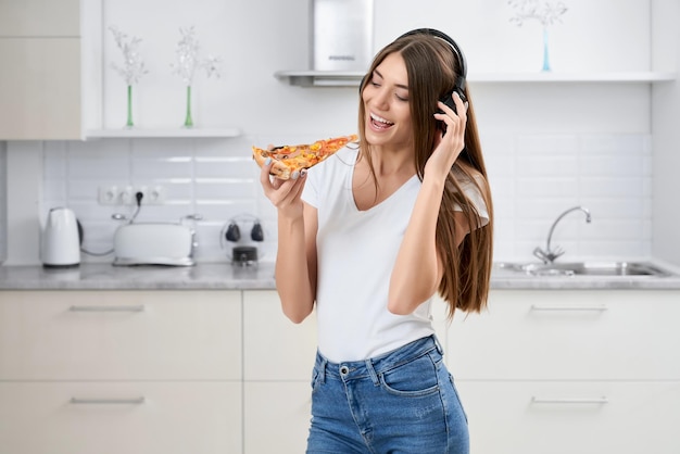 Young woman standing in kitchen and eating pizza