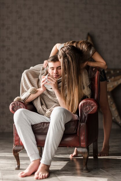 Young woman standing behind the man sitting on armchair holding cup of coffee