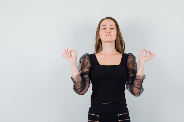 Foto gratuita giovane donna in piedi nel gesto di meditazione in camicetta nera e pantaloni neri e che sembra seria