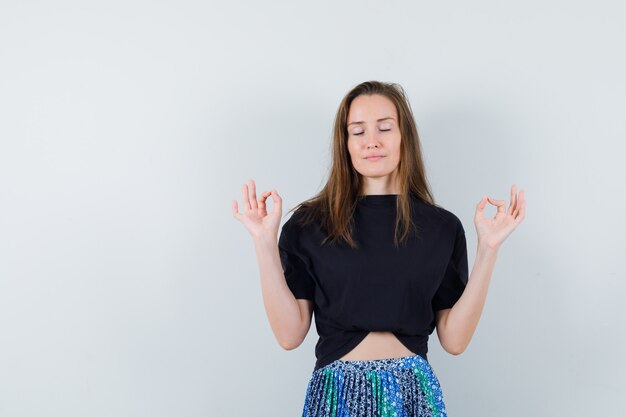 Young woman standing in meditating gesture in black t-shirt and blue skirt and looking calm