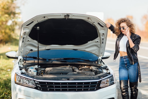 Young woman standing near broken car with popped hood talking on her mobile phone while waiting for help.