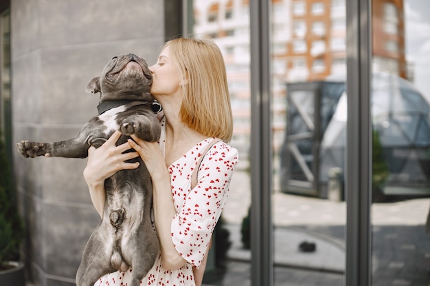 Young woman standing near cafe outdoors holding black French bulldog. Girl wearing black sunglasses and dress