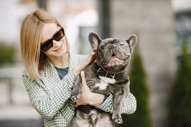 Young woman standing near cafe outdoors holding black French bulldog. Girl wearing black sunglasses, shorts and grey jacket