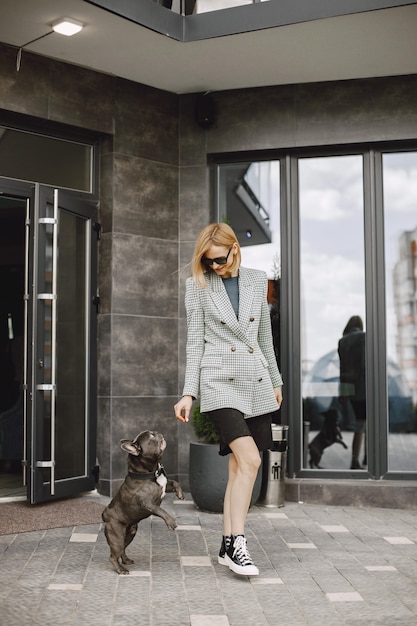 Young woman standing near cafe outdoors with black French bulldog. Girl wearing black sunglasses, shorts and grey jacket