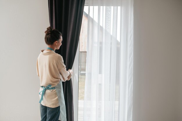 Young woman standing near the widnow and fixing the curtains