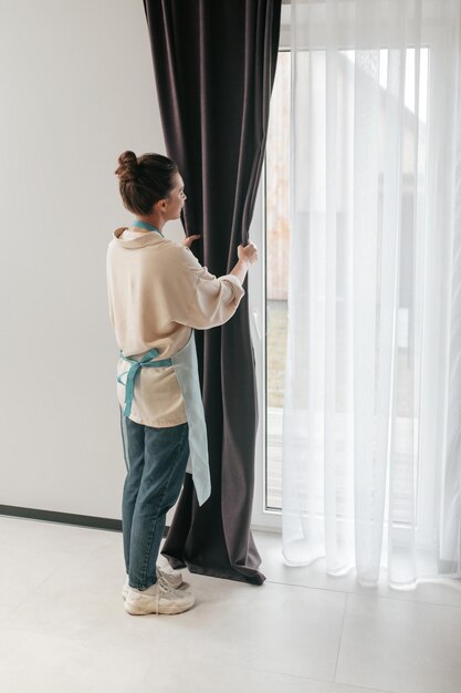Молодая женщина стоит возле окна и чинит шторы