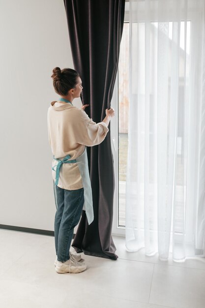 Young woman standing near the widnow and fixing the curtains