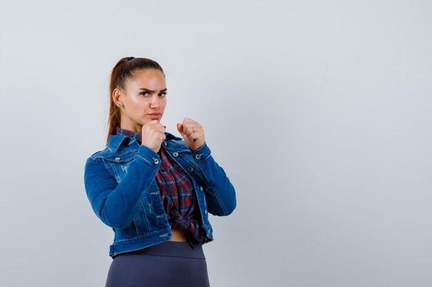 Young woman standing sideways in fight pose in checkered shirt, jean jacket and looking serious.