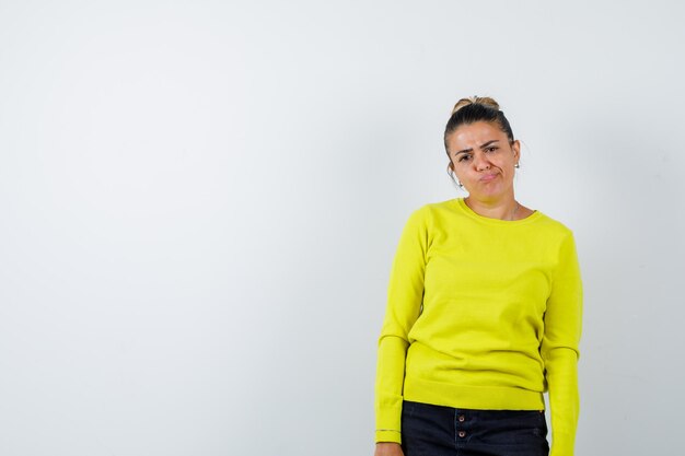 Young woman standing straight, grimacing and posing at camera in yellow sweater and black pants and looking serious