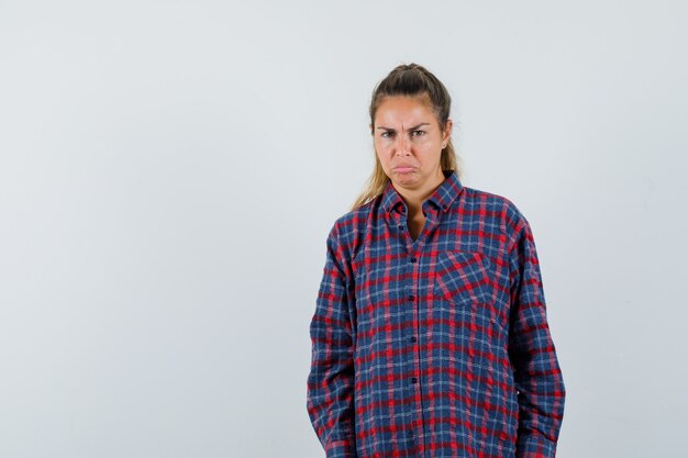 Young woman standing straight, grimacing and posing at front in checked shirt and looking displeased