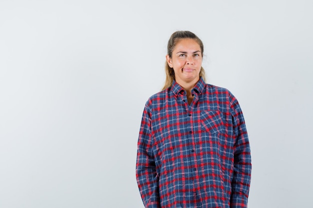 Young woman standing straight, grimacing and posing at front in checked shirt and looking pretty