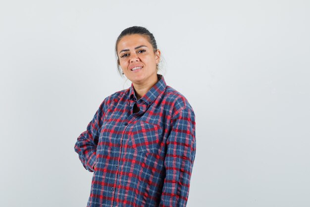 Young woman standing straight and putting hand on waist in checked shirt and looking pretty , front view.