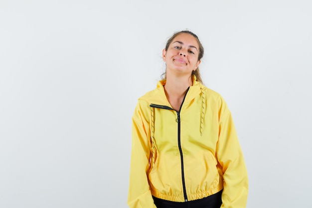 Young woman standing straight, smiling and posing at camera in yellow bomber jacket and black pants and looking cute