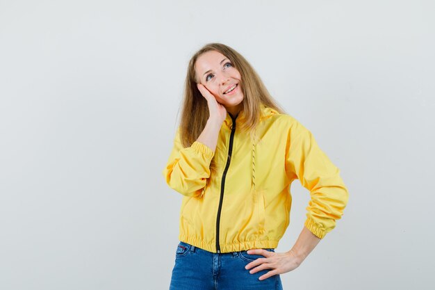 Молодая женщина, стоящая в мысленном жесте, держа руку на талии в желтой куртке-бомбардировщике и синих джинсах, и выглядит оптимистично. передний план.