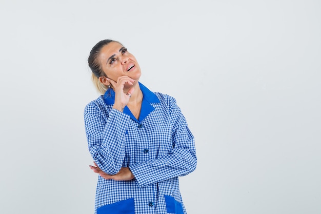 Young woman standing in thinking pose in blue gingham pajama shirt and looking pensive , front view.