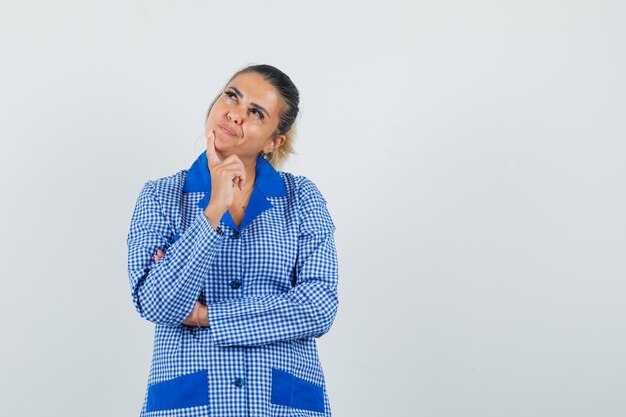 Young woman standing in thinking pose in blue gingham pajama shirt and looking pensive , front view.