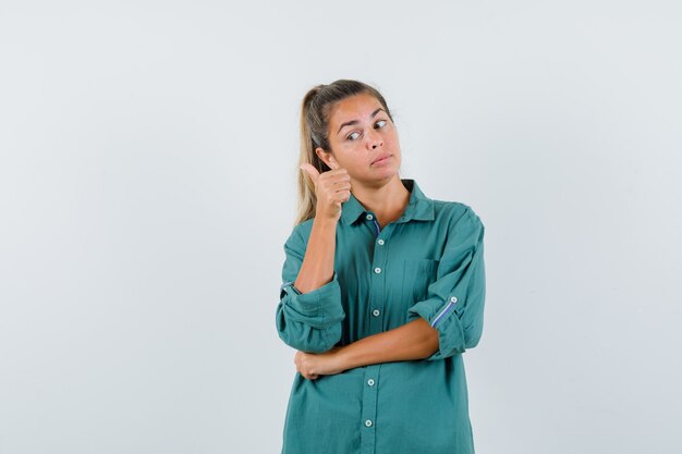 Young woman standing in thinking pose and pointing index finger left in green blouse and looking pensive