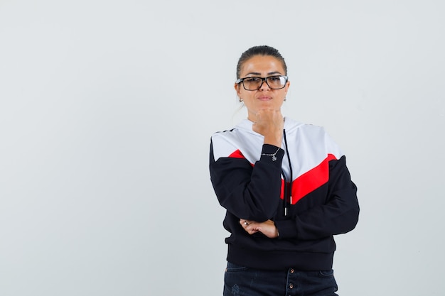 Giovane donna in piedi nella posa di pensiero, mettendo la mano sotto il mento in maglione, jeans neri e occhiali e sembra carina. vista frontale.