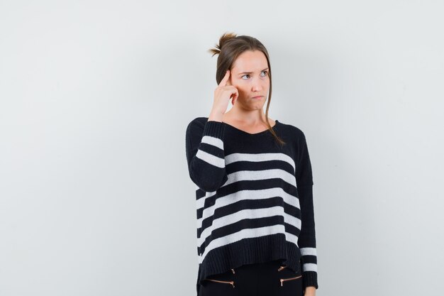 Young woman standing in thinking pose in striped knitwear and black pants and looking pensive
