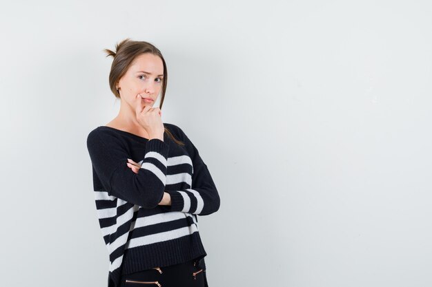 Young woman standing in thinking pose in striped knitwear and black pants and looking pensive