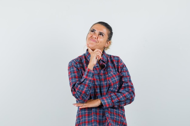 Young woman standing in thinking pose while putting index finger on chin in checked shirt and looking pensive , front view.