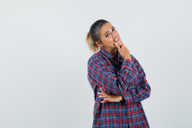 Young woman standing in thinking pose while putting index finger on chin in checked shirt and looking pensive , front view.