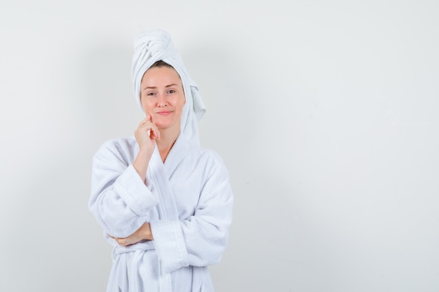 Young woman standing in thinking pose in white bathrobe, towel and looking optimistic. front view.