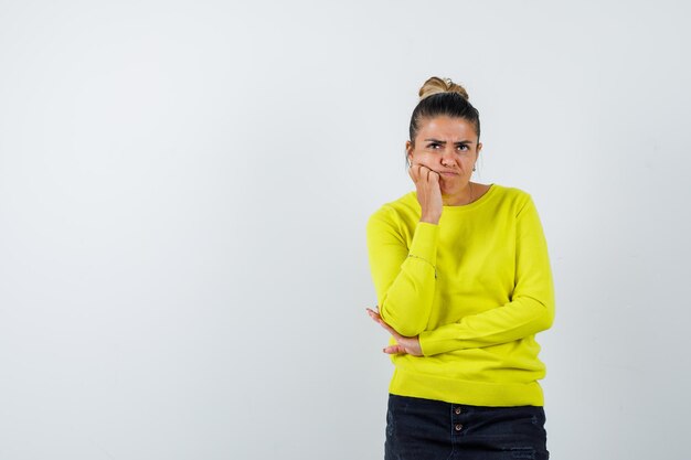 Young woman standing in thinking pose in yellow sweater and black pants and looking pensive