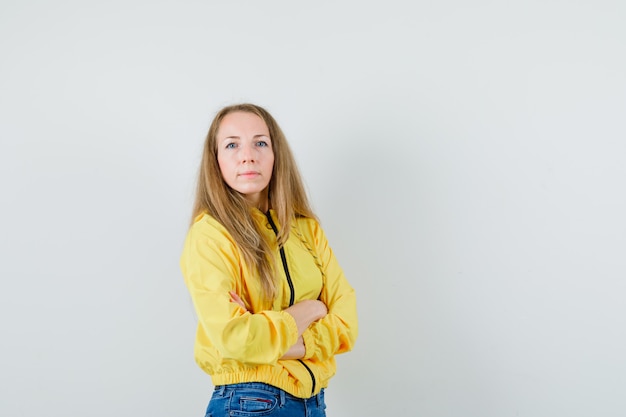 Young woman standing with crossed arms,freezing from cold in yellow bomber jacket and blue jean and looking confident. front view.