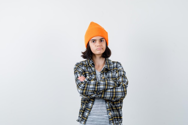 Young woman standing with crossed arms in orange hat checkered shirt looking upset